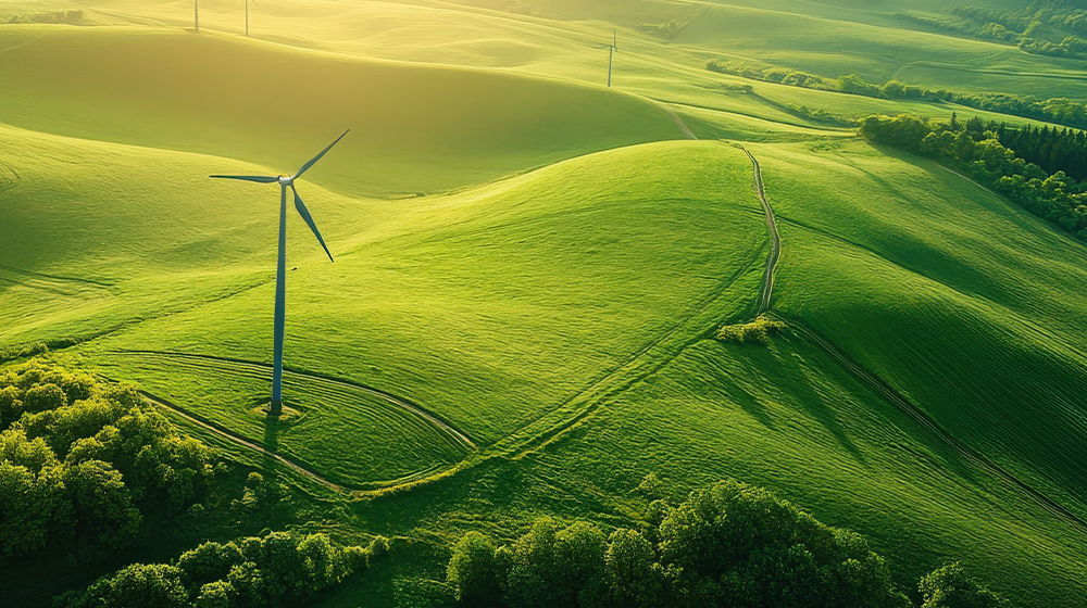 Sanfte grüne Hügel mit einem Windrad im Vordergrund und weiteren Anlagen in der Ferne. Warmes Licht fällt über die Landschaft mit Wegen und Baumreihen.
