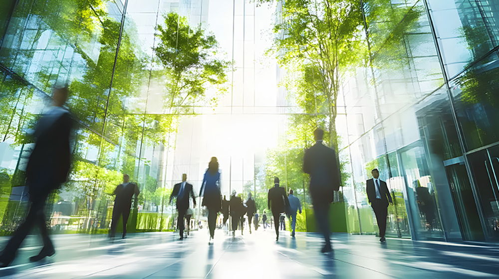 Menschen gehen durch ein modernes, lichtdurchflutetes Gebäude mit Glasfassaden und Bäumen im Innenbereich. Die Szene wirkt geschäftig und urban.