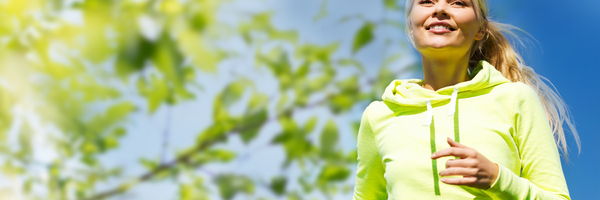 Eine Frau joggt im Freien unter Bäumen und lächelt in die Kamera. Die Szene ist hell und von Sonnenlicht geprägt.