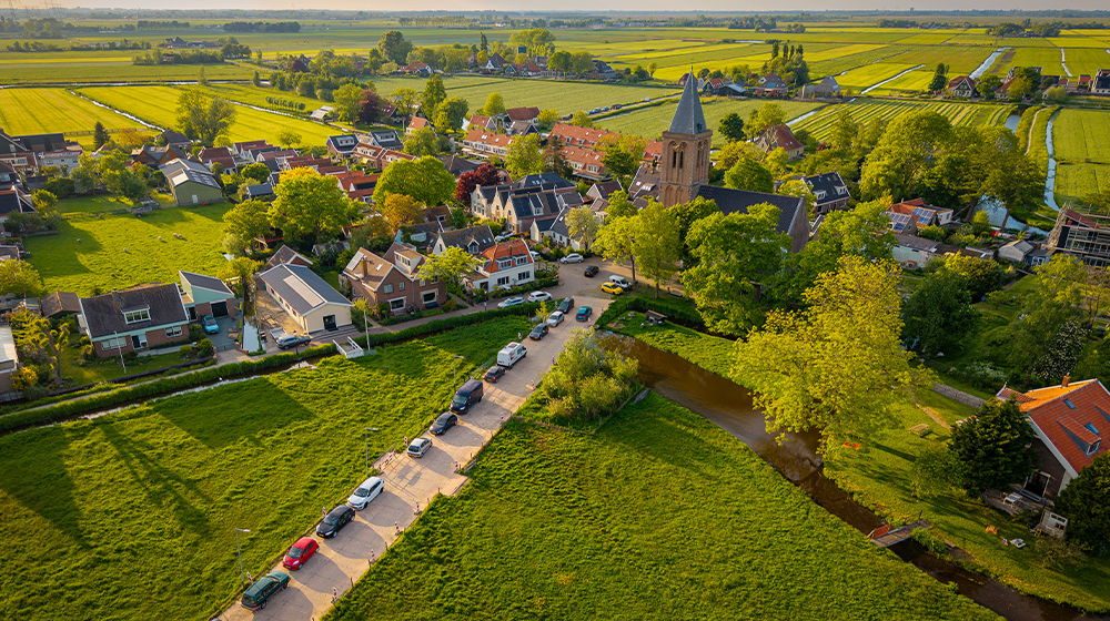 Ein kleines Dorf mit Häusern, Kirche und Straßen liegt eingebettet in eine grüne Landschaft mit Feldern. Eine Straße mit parkenden Autos führt durch die Szene.
